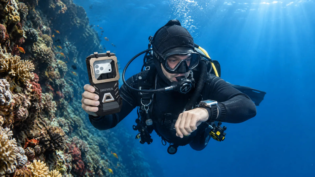 Scuba diver beside a reef wall checking a wrist dive computer while holding a SeaTouch 6 Pro housing with its touchscreen facing him