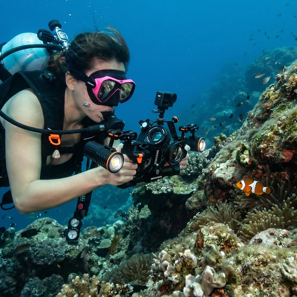 A scuba diver photographing a clownfish on a reef with a DIVEVOLK SeaTouch rig, an action camera mounted on the overhead extension bar