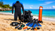 A complete set of scuba diving equipment laid out on a sandy beach, including a wetsuit, BCD, fins, mask, snorkel, dive computer, and an orange surface marker buoy against a tropical ocean background.