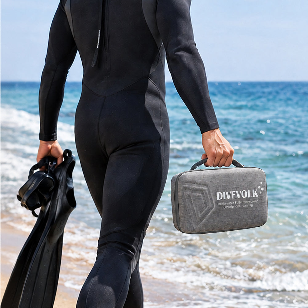 Diver in wetsuit walking into ocean — carrying DIVEVOLK grey travel case in one hand and dive fins in the other, beach dive trip ready.